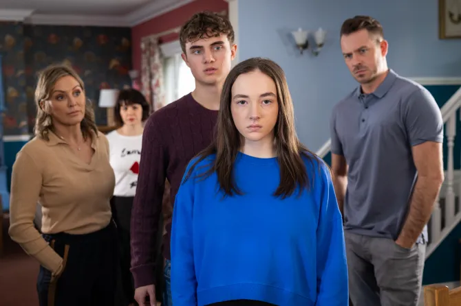 A gril with long dark hair and a blue jumper is looking solemnly into the camera. A boy of the same age is standing behind looking intently at her. Three other adults are further back looking at both of them.  