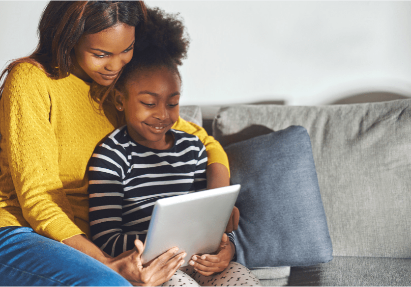 Black woman in a yellow jumper and young black girl stripped top sat on a grey sofa, smiling and looking at a tablet computer.