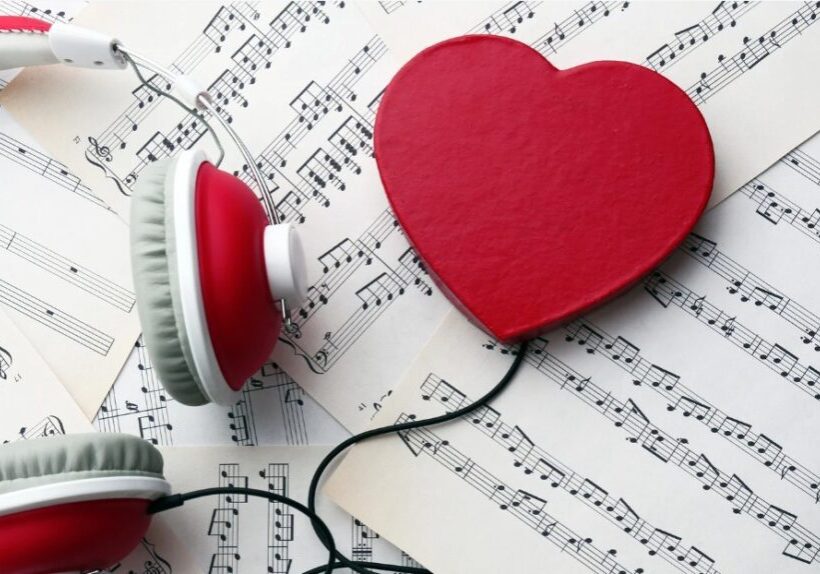 Several sheets of music are scattered overlapping one another. A pair of white and red headphones is on the left hand side. A red wire coming from one of the headphone ears is attached to a red heart.